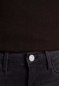 Gros plan sur un pull noir côtelé rentré dans un jean en denim noir taille haute avec un bouton argenté marqué « FRAME ».