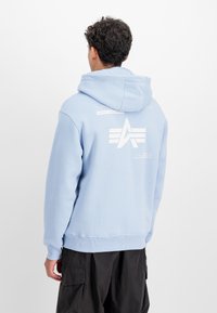 Puha anyagból készült világoskék kapucnis pulóver, nagy fehér logóval a hátsó részen és nyomtatott szöveges díszítésekkel. Bordázott mandzsetták és alsó szegély.