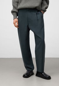 Pantaloni verde scuro con una vestibilità ampia, realizzati in un tessuto leggero. Presentano pieghe frontali e una texture liscia, abbinati a scarpe nere senza lacci.