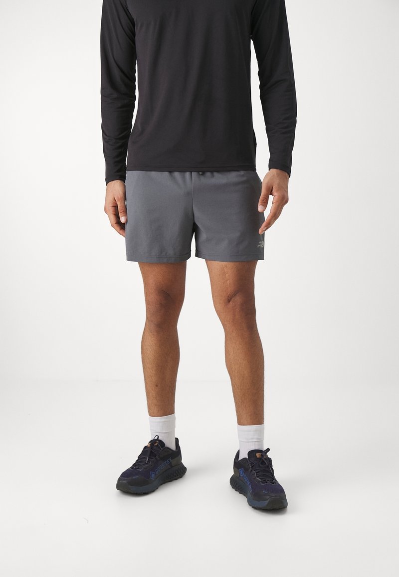 Homme portant un haut à manches longues noir, un short gris, des chaussettes blanches et des chaussures de course noires, debout sur un fond blanc uni.