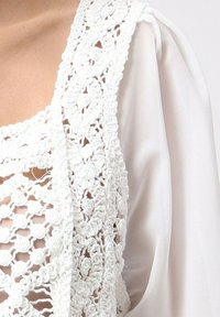 Un tissu en crochet blanc forme un revêtement texturé avec des motifs floraux, superposé sur un matériau blanc lisse et transparent.