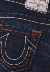 Mörkblå denimjeans med en stor sydd ficka med en distinkt logotyp. Sömmen är i bruna och gräddfärgade nyanser med en mjuk textur.