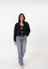Cárdigan negro de punto con un profundo escote en V y tres grandes botones dorados. Lleva puesto sobre una blusa negra y combinado con jeans de cintura alta de color azul claro.