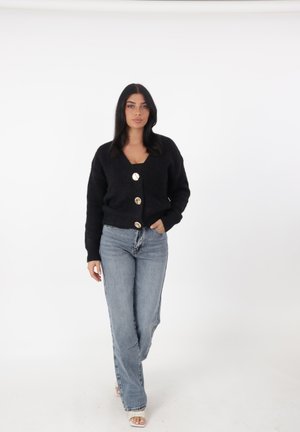 Cardigan nero a maglia con scollatura a V profonda e tre grandi bottoni dorati. Indossato sopra una maglietta nera e abbinato a jeans chiari a vita alta.