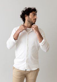 Cămașă albă, cu butoni, dintr-un material moale, având un guler clasic și mâneci lungi, asortată cu pantaloni bej deschis. Fundal neutru.