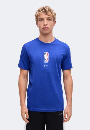 Jeune homme aux cheveux blonds courts, portant un t-shirt bleu NBA et un short noir avec des logos Nike, debout devant un fond clair uni.