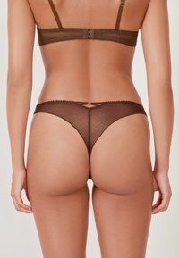 Tanga de encaje marrón transparente con patrón de lunares, presenta un borde ondulado y un lazo en el centro, diseñado con una cobertura mínima.