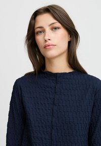 Blouse texturée bleu marine avec un motif géométrique subtil, dotée d'un col rond et d'une fermeture à boutons. Le tissu semble léger et structuré.