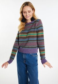 Texturiertes lila Sweatshirt mit mehrfarbigen horizontalen Streifen, versehen mit grünen, blauen und braunen Akzenten, kombiniert mit blauen weit geschnittenen Jeans.