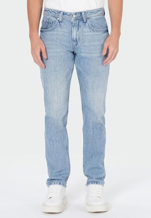 Homme portant un jean droit bleu clair associé à des baskets blanches et une chemise blanche, debout devant un fond uni.