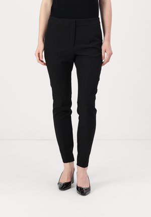 Frau, die enge schwarze Hosen und schwarze Lackleder-High Heels trägt, steht vor einem schlichten weißen Hintergrund.