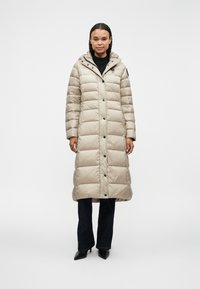 Cappotto imbottito beige con cappuccio, caratterizzato da un design trapuntato, bottoni a scatto e tasche laterali. Abbinato a jeans scuri e scarpe con tacco.