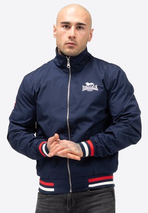 Giacca bomber blu navy con colletto alto, zip frontale argento, polsini a strisce rosse e bianche e logo sul lato sinistro del petto.