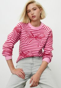 Pull rose avec des rayures rouges et des motifs de nœuds, poignets et encolure côtelés. Porté avec un jean gris clair, mettant en avant une coupe décontractée.