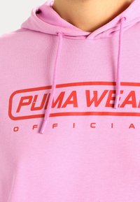 Felpa rosa con cappuccio dotata di cordino e grande logo rosso "PUMA WEAR" sulla parte anteriore. Texture liscia e design casual.