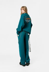 Sudadera de gran tamaño en color azul verdoso con un gran estampado en la parte posterior, combinada con pantalones holgados a juego. Presenta un detalle de cadena y un tejido texturizado.