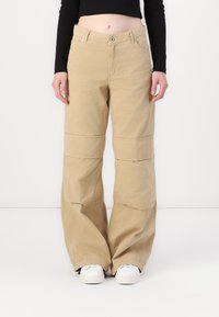 Pantaloni beige a gamba larga in cotone, dotati di zip, tasche frontali e dettagli di cucitura a contrasto; abbinati a sneakers bianche.