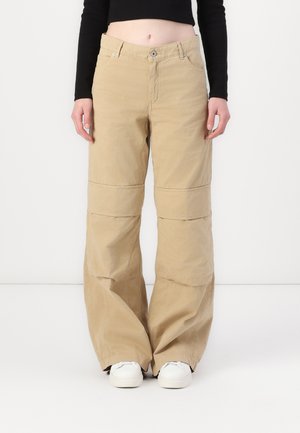 Pantaloni beige a gamba larga in cotone, dotati di zip, tasche frontali e dettagli di cucitura a contrasto; abbinati a sneakers bianche.