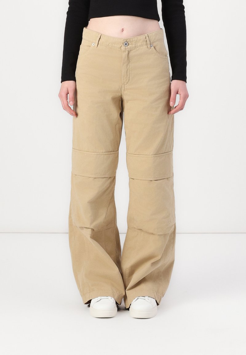 Pantaloni beige a gamba larga in cotone, dotati di zip, tasche frontali e dettagli di cucitura a contrasto; abbinati a sneakers bianche.