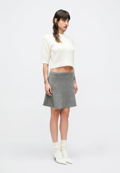 Biały krótki sweter, szara rozkloszowana spódnica, oba wykonane z miękkiej tkaniny. Modelka ma na sobie białe, szpiczaste botki i kremowe skarpetki. Prosty design, neutralne kolory.