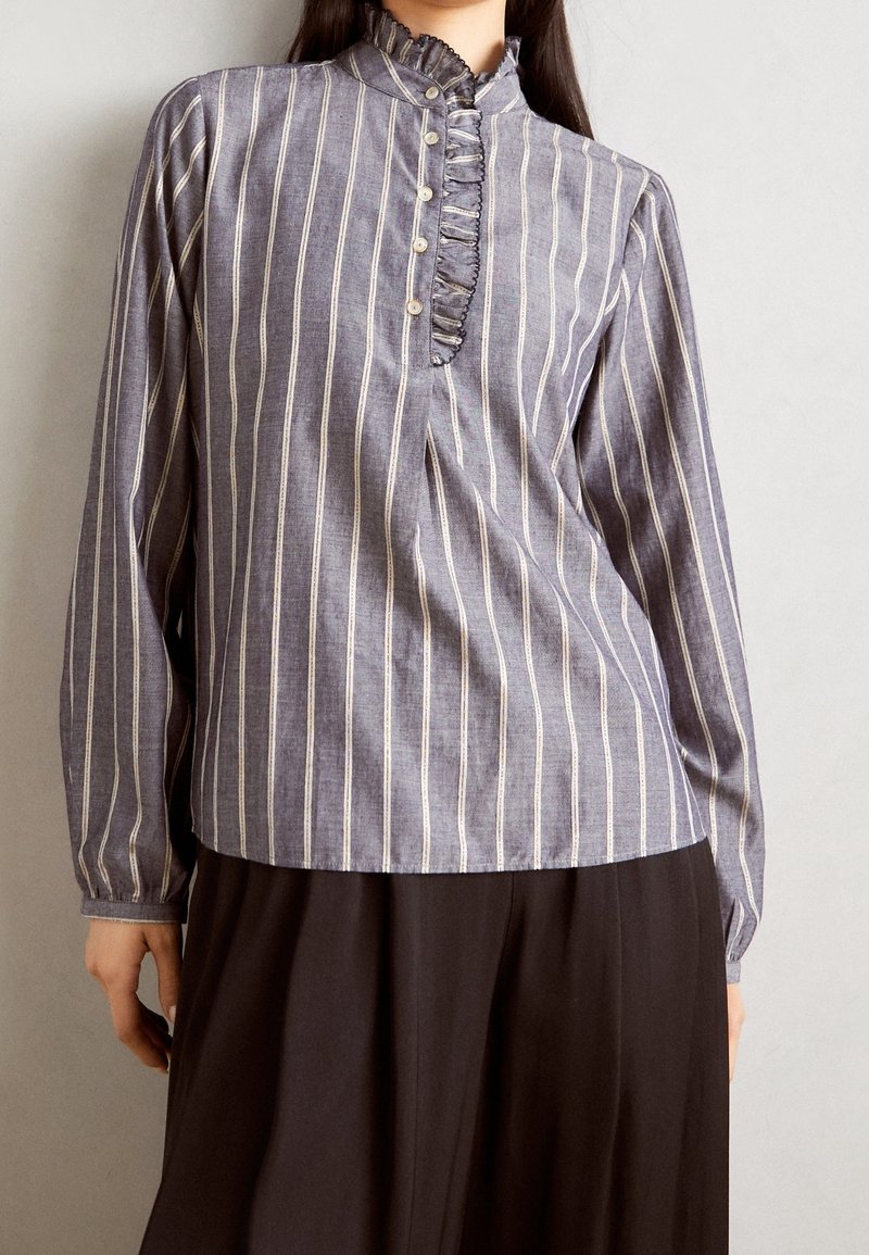Femme portant un chemisier à manches longues rayé verticalement gris et blanc avec col à volants et patte de boutonnage, assorti à une jupe noire.