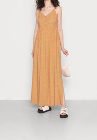 Robe maxi fleurie orange à fines bretelles, avec des boutons sur le devant et une jupe évasée. Associée à des sandales plates crème et un petit sac à main.