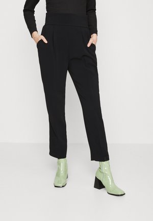Zwarte op maat gemaakte broek met een hoge tailleband, voorzien van plooien en zijzakken. Gecombineerd met groene laarzen tot de enkel van patentleer met blokhakken.