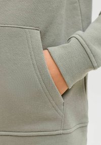 Detaj svetlo zelenega puloverja z bočno žepom in manšetami. Tkanina je teksturiran bombaž s vidnimi šivi na robovih.