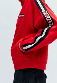 Červená mikina s černými a bílými pruhy podél rukávů. Má vysoký límec, přední kapsu na zip a vyšívané logo na hrudi.