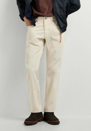 Mann trägt cremefarbene gerade geschnittene Hose, dunkelbraune Stiefel, ein braunes Hemd und eine marineblaue Reißverschlussjacke mit den Händen in den Taschen.