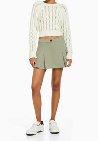 Cremfarbener Strickpullover mit Lochmuster, kombiniert mit einem plissierten olivgrünen Rock mit Seitentaschen und Knopfverschluss. Weiße Sneakers.