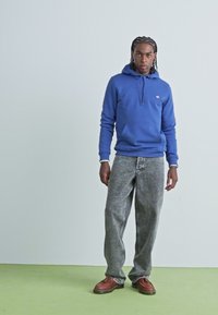Sweatshirt bleu à capuche avec poche avant, associé à un jean gris ample à motifs et des chaussures marron, le tout sur un fond clair.