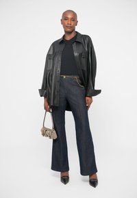 Chemise en cuir noir, haut ajusté noir, pantalon en denim foncé évasé avec détail en chaîne dorée, sac à main gris et chaussures noires à bout pointu.