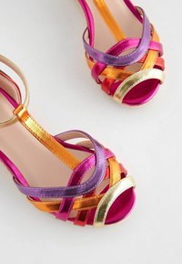Metallicsandaletter med remme og åben tå i pink, lilla, orange, rød og guld med ankelremme på en hvid baggrund.