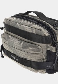 Grau camouflierter Beutel mit gepolsterter Textur, doppelten Reißverschlüssen und schwarzen Gurten. Verfügt über ein auffälliges schwarzes Markenlogo an der Seite.