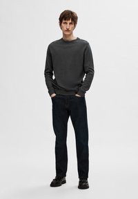 Dark grey sweater with a round neckline and ribbed cuffs, paired with dark denim jeans and black shoes, standing against a light background.