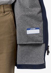 Veste K-Way avec un extérieur bleu marine et un intérieur en polaire grise. Comprend une étiquette avec un nom bleu et un détail de fermeture éclair au bas.