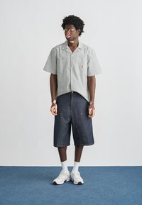 Ριγέ πουκάμισο με κοντά μανίκια και τσέπη στο στήθος, σε συνδυασμό με σκούρα τζιν σορτς που διαθέτουν εμπρόσθιες τσέπες και χρυστά ράμματα. Λευκά αθλητικά παπούτσια και κάλτσες.