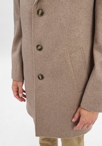 Beige Wollmantel mit drei braunen Knöpfen und Seitentaschen, getragen über braunen Hosen, mit Händen an den Seiten vor einem weißen Hintergrund.