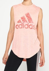 T-shirt sans manches rose clair en tissu doux et texturé, arborant un grand logo Adidas rouge. Ourlet arrondi et coupe décontractée.