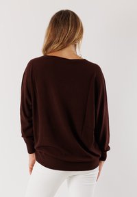 Donna con capelli castano chiaro che indossa un maglione scuro marrone largo e pantaloni bianchi, rivolta con il dorso a un fondo chiaro e uniforme.