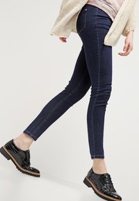 Tmavě modré skinny džíny s hladkým povrchem, s kontrastním prošitím, doplněné lesklými černými oxfordkami a světlým pleteným kardiganem.