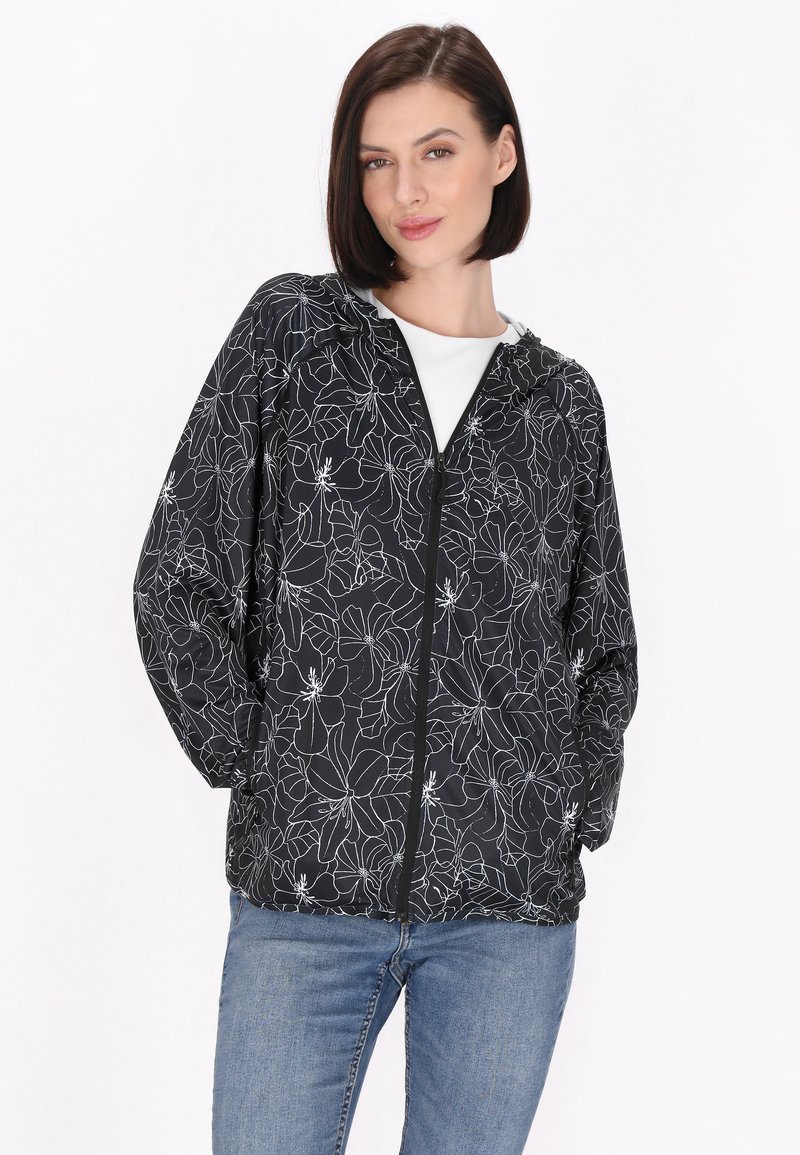 Jachetă neagră cu model floral alb, glugă și închidere cu fermoar. Material ușor, mâneci lungi și croi lejer, purtată cu blugi albaștri.