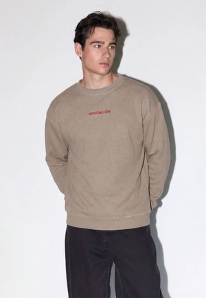 Joven con cabello oscuro que lleva un suéter beige con texto rojo "unsubscribe" y pantalones negros, de pie sobre un fondo blanco.