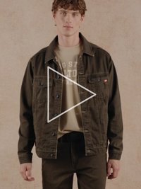 Smeđa traper jakna s crnim gumbima, u kombinaciji s bež majicom s bijelim natpisom. Model također nosi tamne hlače, stojeći na neutralnoj pozadini.