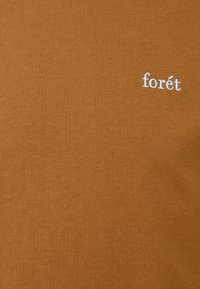 T-shirt marron en tissu de coton doux, avec un petit logo blanc brodé "forét" sur le côté gauche. Texture lisse, design décontracté.