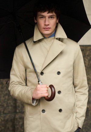 Young man in beige double-breasted trench coat holding a black umbrella with wooden handle, standing against a dark stone background.