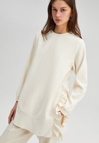 Light cream tunic with long sleeves, soft texture, featuring ruffled side accents and a round neckline, designed for a relaxed fit.