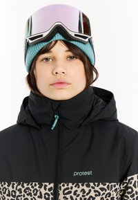 Giacca leopardata nera e beige con cappuccio, caratterizzata da una chiusura zip e dettagli turchesi. Indossata con un berretto in maglia turchese e occhiali da sci.