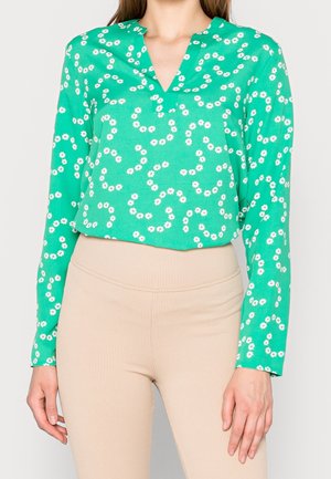 Woman wearing a bright green blouse with tiny white flower prints and beige ribbed high-waist pants, shown from shoulders to hips.